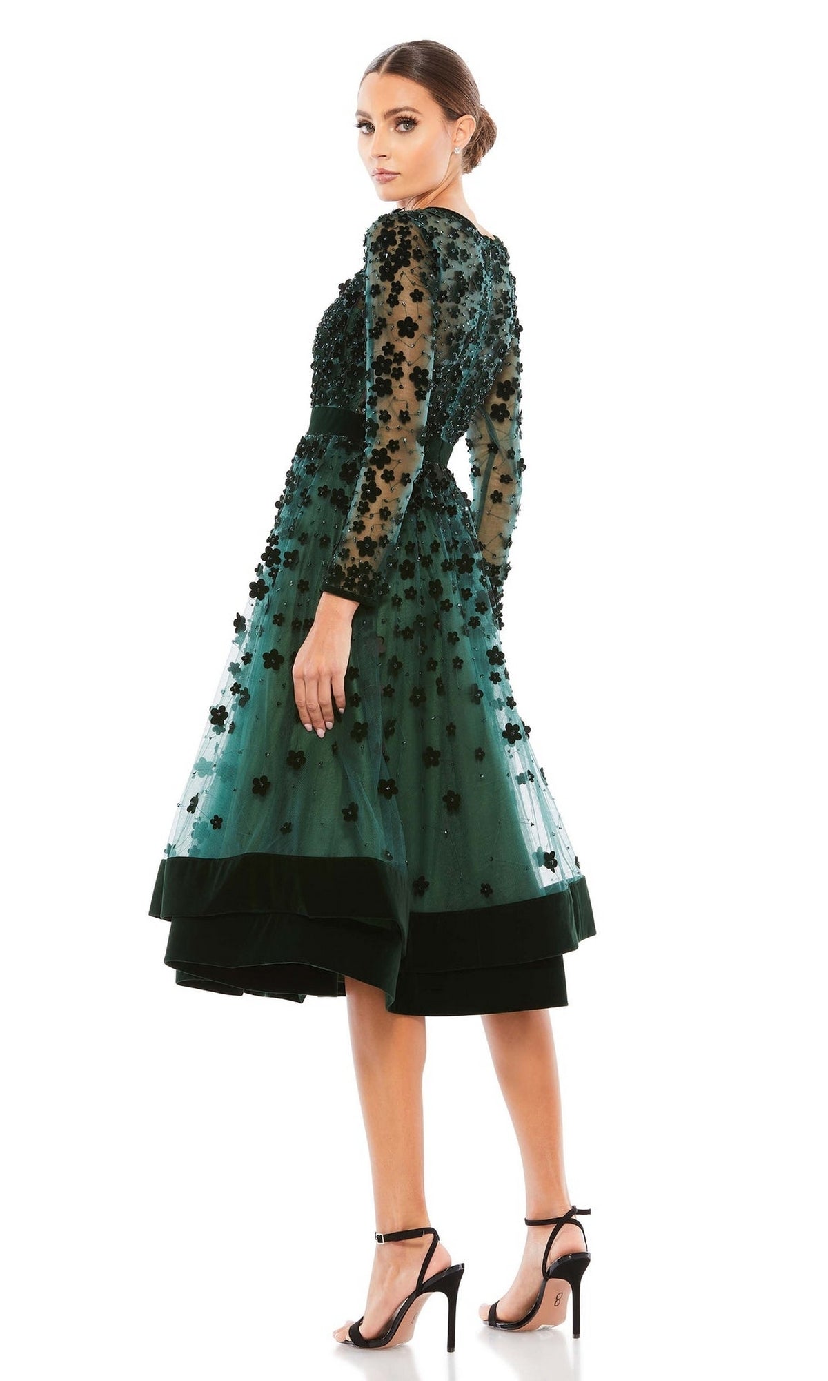 Woman in the Mac Duggal 67007 knee-length wedding-guest dress with green floral embellishments and sheer long sleeves, paired with high-heeled sandals, standing on a white background.